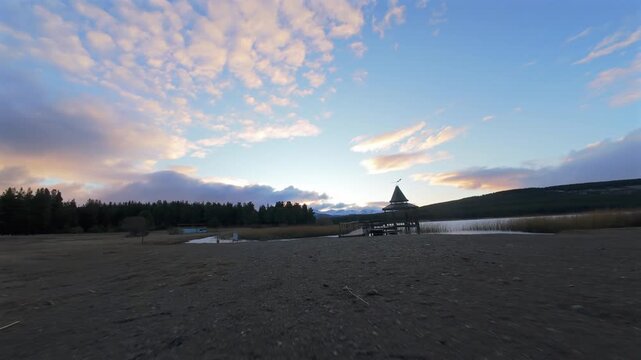 Drone flight past a wooden pier and gazebo on the calm waters of Laguna La Zeta in Esquel, featuring sky reflections.