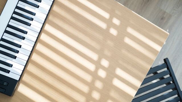 Midi keyboard on a wooden table beside a chair with warm sunlight and window shadows in a peaceful home studio or relaxation space.