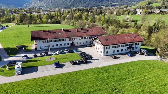 Aerial view of the Nazi Berchtesgaden Chancellery building with red roofs, parked cars, green meadows, and Bavarian Alps in Germany.