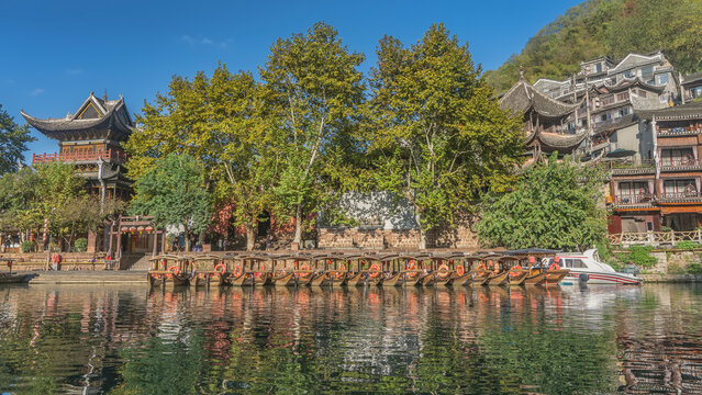 Wooden tourist boats are moored in a row by the river bank. Canopies, lifebuoys on the decks. There are green trees, traditional Chinese buildings on the embankment. The blue sky. China. Fenghuang
 