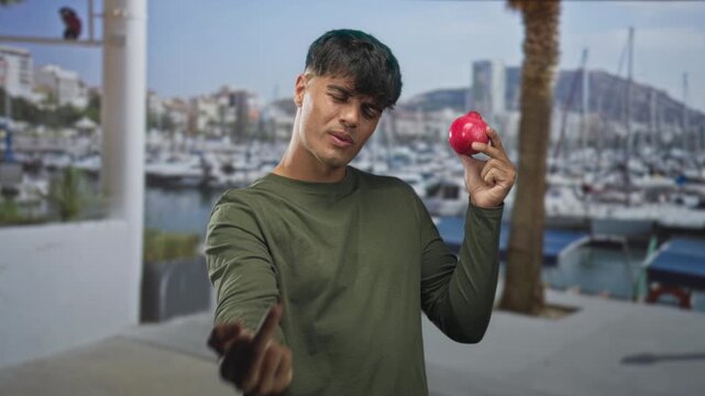 Young hispanic man holding a red apple with puckered lips shows middle finger toward camera in front of building; defiance rebellion.