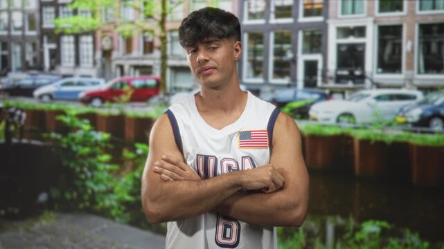 Man in usa basketball jersey with flag patch crossing arms and gesturing with hand on a street; quiet confidence.