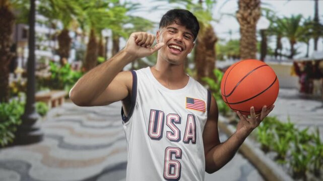 Man holding basketball points thumb to chest on street with palm trees and patterned walkway while wearing usa jersey and smiling; playful confidence.