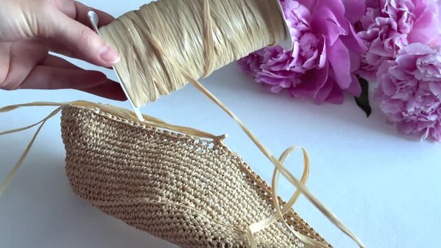 Woman hands holding a spool of natural raffia ribbon over a woven straw bag and pink peonies. DIY craft, handmade bag, summer accessories