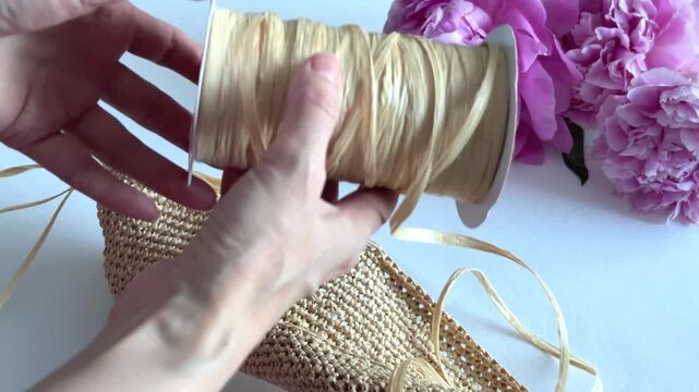 Woman hands holding a spool of natural raffia ribbon over a woven straw bag and pink peonies. DIY craft, handmade bag, summer accessories