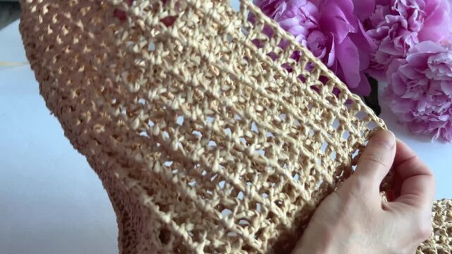 Hands holding handmade crochet bag made from natural raffia fiber with pink peonies in background, craft hobby project display.