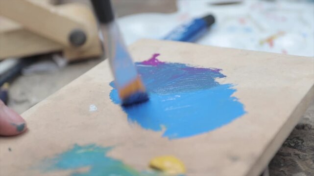 An artist's hand mixes colors with a paintbrush on a palette. The palette is brown, and the colors being mixed are light blue, purple, and yellow, with green gradually appearing.