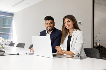 Portrait of two happy multinational colleagues, Hispanic and Indian team members smiling at camera sit at modern office desk with laptop. Teamwork, diversity, leadership, positive corporate workflow