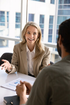 Smiling businesswoman in elegant jacket sitting at conference desk, lead formal talk with client or colleagues, express idea or solution at meeting. Applicant answers questions during job interview