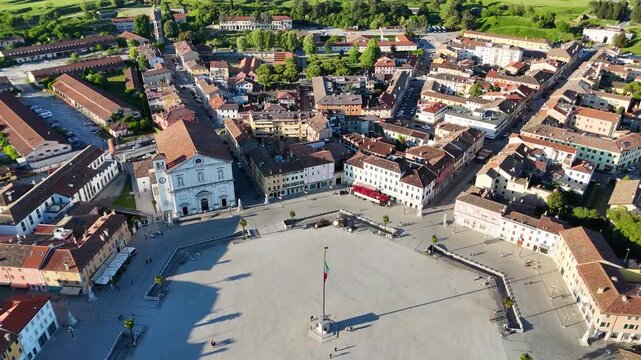 Drone view of Palmanova, Italy, featuring Piazza Grande, a white church facade, terracotta roofs, green countryside, and distant mountains.