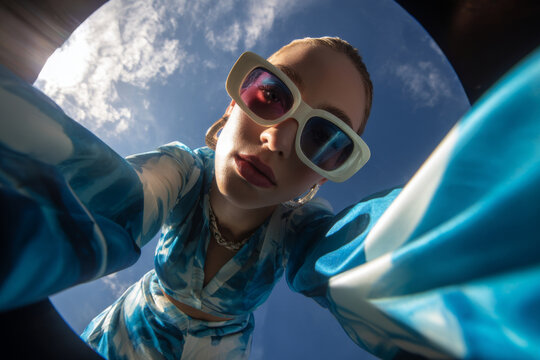 Low angle view of a woman in artistic fashion makeup wearing oversized sunglasses, looking down into the camera against a bright blue sky with clouds