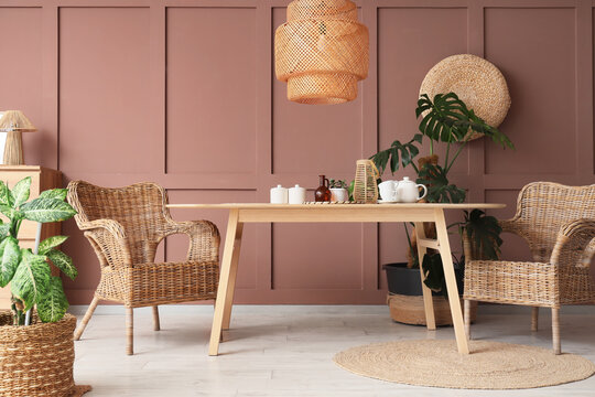 Boho chic dining room interior with wicker furniture, wooden table, and houseplants against a dusty pink wall