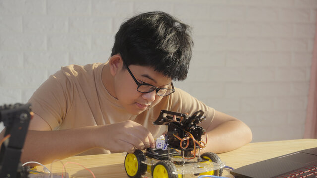 Asian young student assembling a robotics car at home during STEM learning. Concept of coding education, engineering skills, innovation, and hands-on technology training.