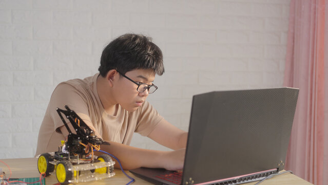 Asian young student using a laptop at home while working on a robotics project with electronic components and robotic devices. Concept of STEM education, coding, and technology learning.