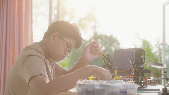 Asian young student wiring a robotics project at home with robotic devices, electronics, and a laptop. Concept of STEM education, engineering skills, innovation, and hands-on technology learning.