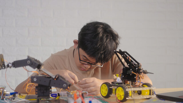 Asian young student wiring a robotics project at home with robotic devices, electronics, and a laptop. Concept of STEM education, engineering skills, innovation, and hands-on technology learning.