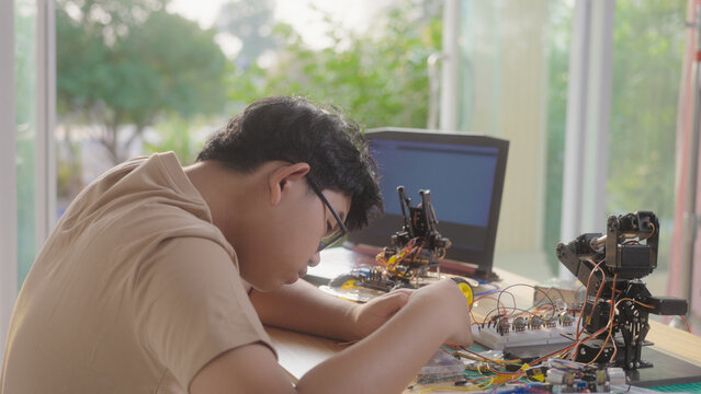 Asian young student building electronic circuits and a robotics project at home with robotic devices and a laptop. Concept of STEM education, engineering skills, and technology learning.