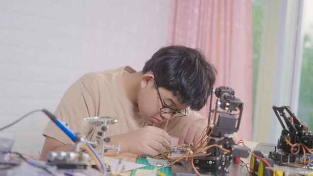Asian young student wiring a robotics project at home with robotic devices, electronics, and a laptop. Concept of STEM education, engineering skills, innovation, and hands-on technology learning.