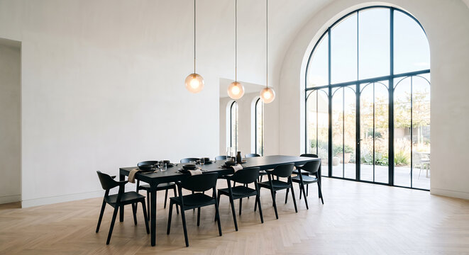 A modern dining room with a long black table set for a meal, surrounded by black chairs. Large arched windows offer a view of a sunny garden, and pendant lights hang from the high ceiling.