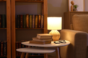 Cozy home interior with bookshelf, reading lamp, stack of books, and eyeglasses on a side table © Pixel-Shot