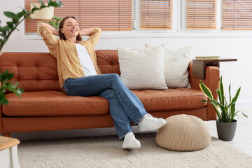Obraz premium Relaxed young woman enjoying leisure time on comfortable sofa in bright living room interior, she is happy and peaceful, with her eyes closed and hands behind her head