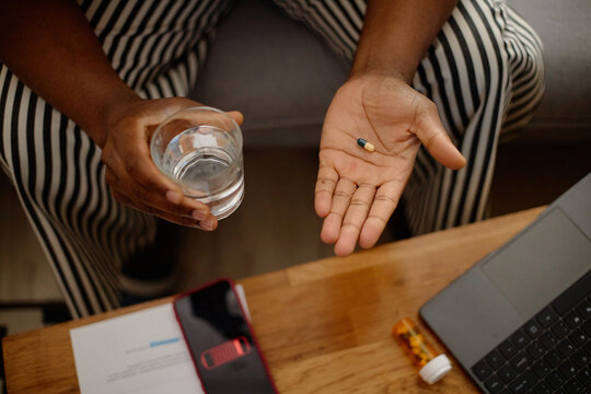 Close up of Black man holding capsule and glass of water near smartphone and laptop, showing screen fatigue or digital addiction at home. Useful for mental health or wellness campaigns