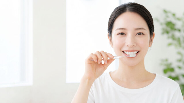 明るく清潔感のある室内で歯ブラシを手にし、笑顔でオーラルケアを行う若い女性の爽やかなライフスタイルイメージ。白い歯と健康的な表情、ナチュラルな日常シーンが印象的で、歯磨き、口腔衛生、デンタルケア、予防習慣、朝のセルフケアやヘルスケア広告に使いやすい親しみのある人物素材