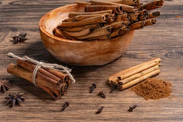Cinnamon sticks in wooden bowl with ground cinnamon powder and various spices scattered on a rustic surface © panadesignteam
