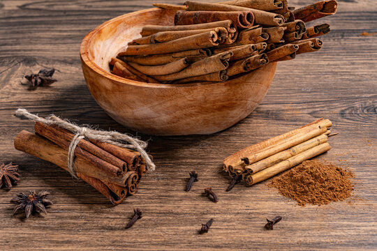 Cinnamon sticks in wooden bowl with ground cinnamon powder and various spices scattered on a rustic surface