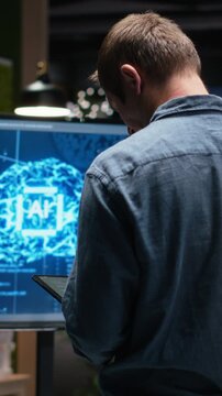 Portrait video of programmer in office using tablet, working to fix bugs in AI system. IT specialist troubleshooting neural network errors with device in tech startup workplace.