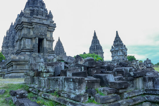 Ancient temple ruins stretch across green grassy terrain at Prambanan Temple. Weathered stone blocks lie scattered in foreground. spires rise against pale sky. Lush tree provides contrast 