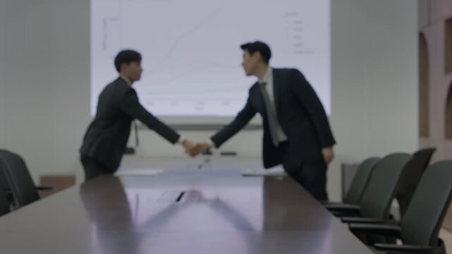 Two businessmen in dark suits shaking hands across a conference table in a boardroom with a projected line chart on the screen behind them.