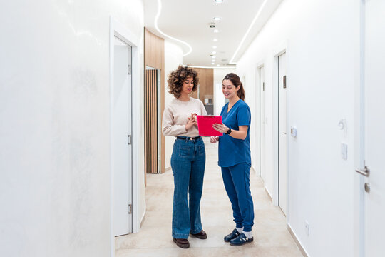 Medical worker in blue scrubs holding pink clipboard while talking to female patient with curly hair in clinic hallway. 