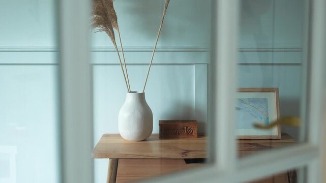 A serene interior scene featuring a vase with dried plants on a wooden table through a doorway.