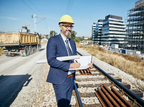 senior businessman architect wearing a protective helmet writing in a notebook and holding a blueprint on the construction site