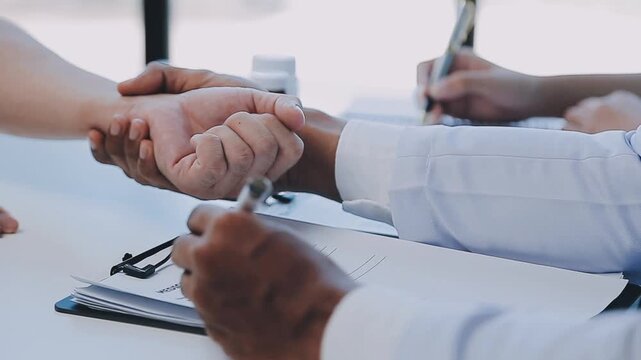 A close-up shot of a doctor's hands checking a patient's pulse on their wrist while another person's hand is visible in the background holding a pen and writing on a clipboard, suggesting a medical