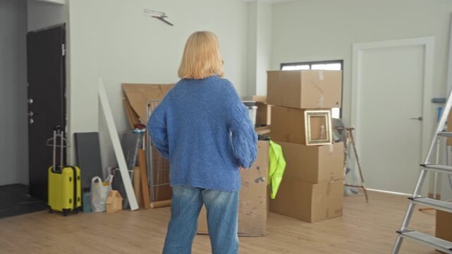 Woman in blue sweater with hands on hips beside stacked moving boxes in building; packing determination.