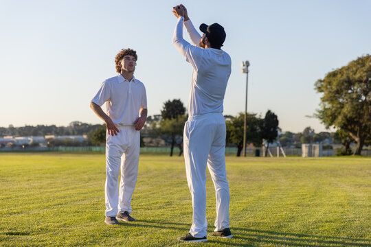Two male cricket players catching ball on grassy suburban cricket ground, wearing white uniforms