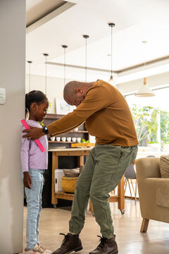 African American dad bending, measuring schoolgirl with pink ruler at wall in kitchen, copy space
