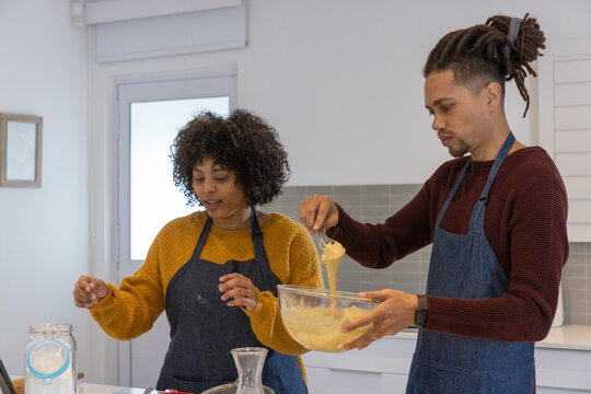 Diverse couple whisking batter together at kitchen island, wearing aprons and holding bowl and jar