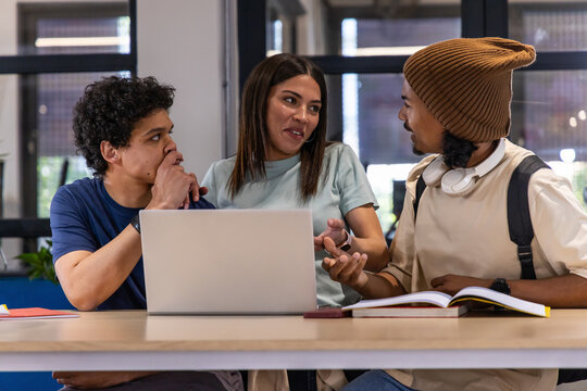 Diverse students sitting closely at table in study, discussing silver laptop and open textbook