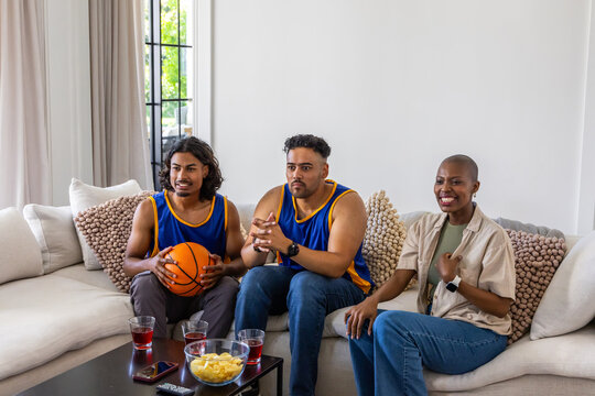 Diverse friends wearing basketball jerseys watching game at home on couch with orange basketball