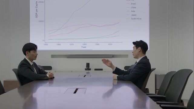 Two businessmen in dark suits gesturing during a discussion at a conference table in a boardroom with a large projected GDP line chart displayed on the screen behind them.