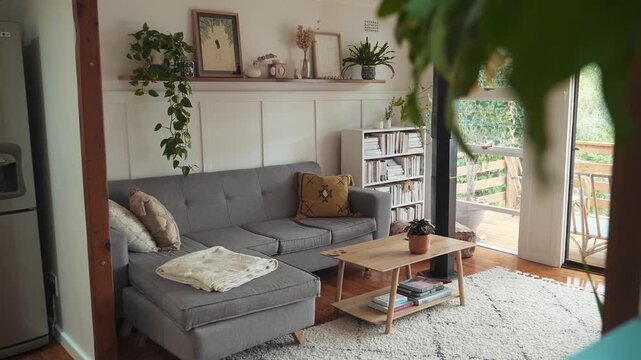 A cozy living room with a gray sofa, wooden coffee table, and plants on a shelf in a house interior with a view.