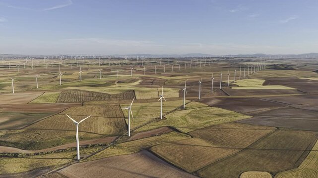 High and wide aerial view of large wind farm in Spain. HDR drone footage flying forward over rows of wind turbines turning in the wind spread out across a patchwork of fields in Zaragoza, Spain.