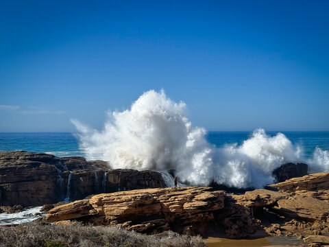 Unknown person standing on a rocky outcrop, dwarfed by the immense wave crashing behind. Huge plume of white foam highlights sheer power of the ocean, while brown cliffs add a sense of rugged beauty.
