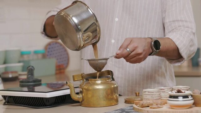 Man pouring coffee from metal pot through strainer into brass coffee pot on kitchen counter with various baking items