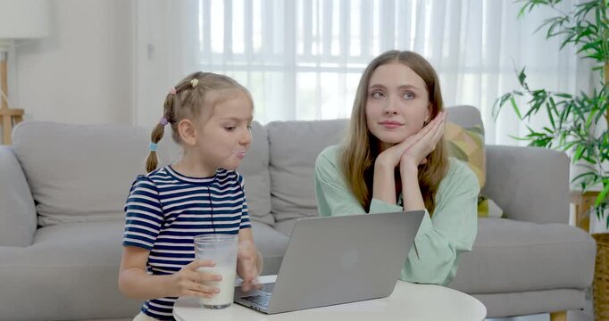 Caucasian single mother listens as preschool daughter talks beside laptop and milk glass, supporting communication, online lesson, gentle guidance, daily childcare, cozy home learning routine.