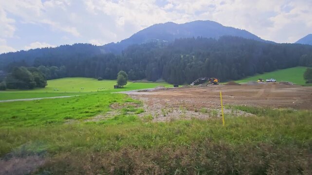 Railway travel view of open green valley, mountain forest, roadside fence, and rural buildings, filmed from a train window for scenic countryside, tourism, and journey videos stock footage.