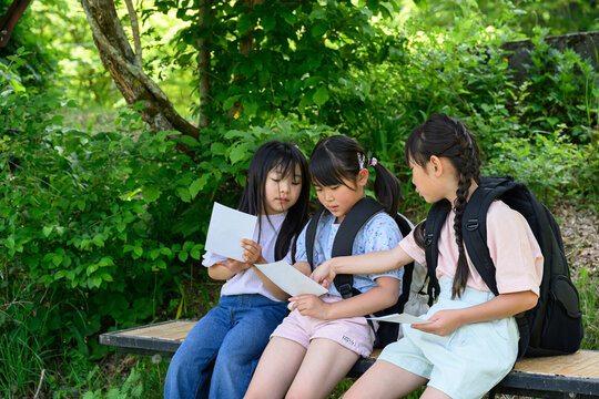 新緑の公園のベンチに座って地図を見ながら相談するリュックを背負った女子小学生たち